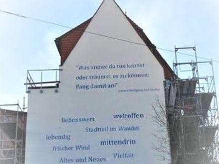 Hauswand des Gebäudes der Goethestraße 60. Um das Gebäude wurden Baugerüste aufgebaut. Die Hauswand wird von blauer Schrift geziert. Dort stehen Worte, wie „weltoffen“, „zukunftsorientiert“, usw.