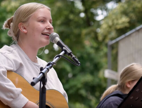 Junge, blonde Frau singt in ein Mikrofon. Gleichzeitig spielt sie Gitarre. Im Hintergrund sieht man Baumkronen.