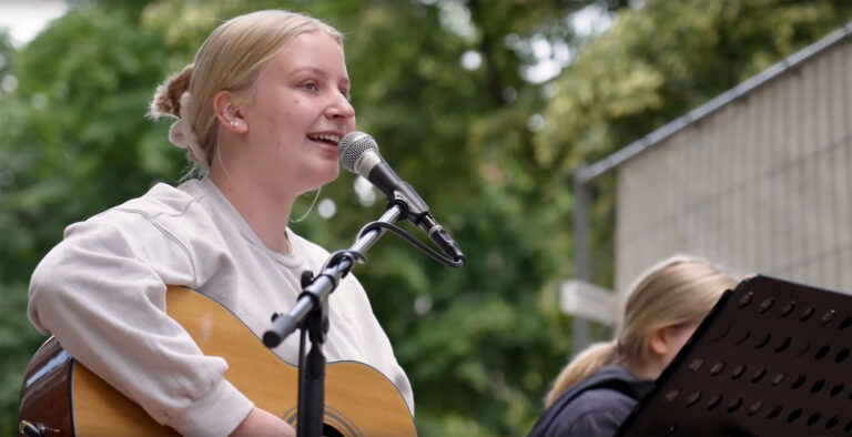 Junge, blonde Frau singt in ein Mikrofon. Gleichzeitig spielt sie Gitarre. Im Hintergrund sieht man Baumkronen.
