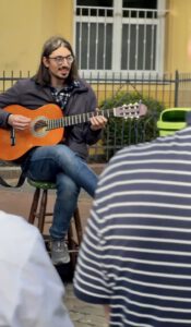 Musiker sitzt mit einer Gitarre auf einem Hocker und lächelt. die Zuschauenden an. Im Hintergrund sieht man eine gelbe Hauswand mit einem Zaun davor.