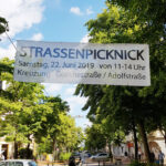 In den Bäumen der Goethestraße in Bremerhaven Lehe wurde ein großes weißes Banner gespannt auf dem das Straßenpicknick in schwarzen Buchstaben beworben wird. Auf der Straße sieht man Menschen, die Tische und Bänke aufbauen.