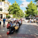 Viele Menschen sitzen an den Tischen und Bänken für ein Straßenpicknick auf der Kreuzung einer verkehrsberuhigten Straße.
