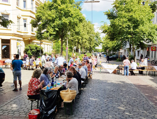Viele Menschen sitzen an den Tischen und Bänken für ein Straßenpicknick auf der Kreuzung einer verkehrsberuhigten Straße.