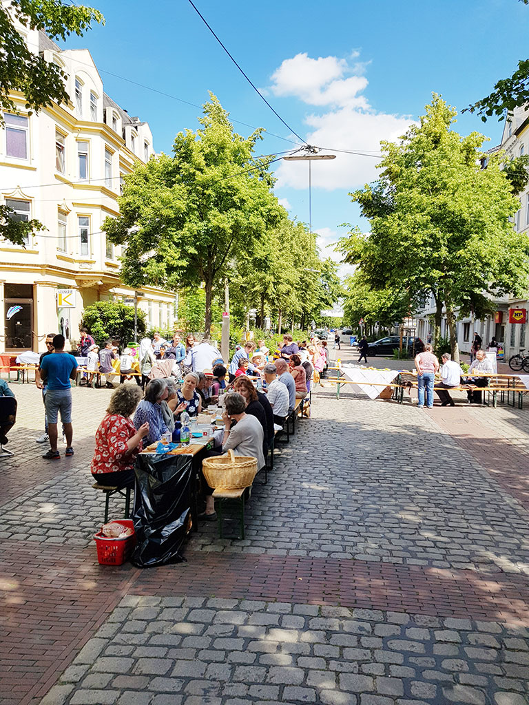Viele Menschen sitzen an den Tischen und Bänken für ein Straßenpicknick auf der Kreuzung einer verkehrsberuhigten Straße.
