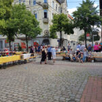 Menschen stehen, sitzen und reden beim Straßenpicknick. Mehrere Bierzeltgarnituren wurden auf der Kreuzung angeordnet und mit Tischdecken ausgestattet.