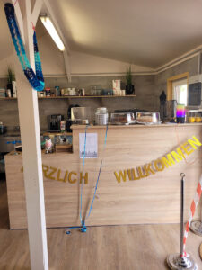 Eröffnung des Nachbarschaftscafés „Bunte Bude“. Der Innenraum hat eine helle Holzverkleidung. An der Theke ist eine Girlande mit den goldenen Buchstaben „Herzlich Willkommen“ befestigt.