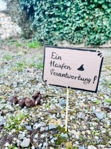 Aus Pappe gebasteltes Schild mit der Aufschrift „Ein Haufen Verantwortung!“ wurde mit einem kleinen Holzstab auf einem Gehweg neben Hundekot platziert.