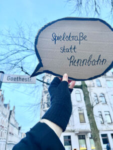 Aus Pappe ausgeschnittene Sprechblase mit der Aufschrift „Spielstraße statt Rennbahn“ wird von einer Person hochgehalten. Im Hintergrundsieht man das Straßenschild „Goethestr.“ und Gebäude.