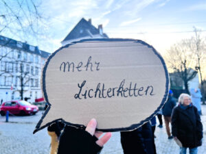 Aus Pappe ausgeschnittene Sprechblase mit der Aufschrift „mehr Lichterketten“ wird von einer Person hochgehalten. Im Hintergrund ist ein Teil des Goetheviertels zu sehen.