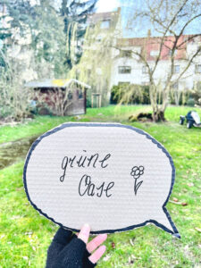 Aus Pappe ausgeschnittene Sprechblase mit der Aufschrift „grüne Oase“ wird von einer Person hochgehalten. Im Hintergrund ist eine grüne Wiese, ein Gartenhaus, ein Baum und Häuser im Goetheviertel zu sehen.