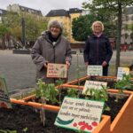 Zwei Anwohnerinnen halten selbst gebastelte, mehrsprachige Schilder mit Beschriftungen für die neu bepflanzten Hochbeete auf dem Leher Pausenhof hoch. Auf den Schildern steht zum Beispiel „Lass mich in Ruhe wachsen.“ oder „Bitte pflege mich, ich belohne dich mit meinen Früchten.“
