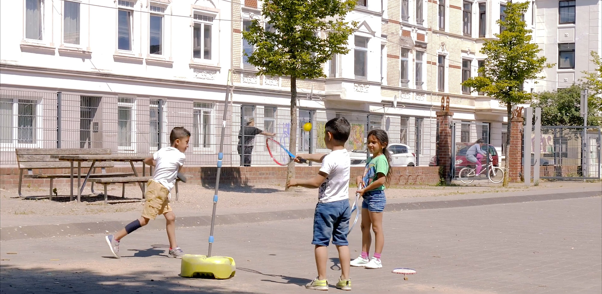 Drei Kinder spielen auf dem Leher Pausenhof. Im Hintergrund fährt ein weiteres Kind auf einem Fahrrad durch das offene Tor auf den Leher Pausenhof.