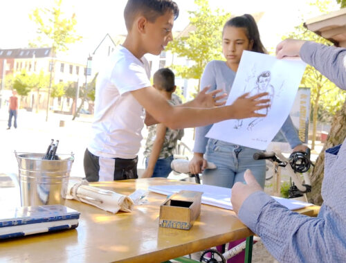 Ein älterer Mann sitzt an einem Kinder-Maltisch auf einem öffentlichen Spielplatz im Goetheviertel. Im Hintergrund sind mehrere Kinder zu sehen. Der Mann übergibt einem Jungen eine Zeichnung.