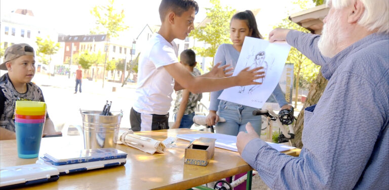 Ein älterer Mann sitzt an einem Kinder-Maltisch auf einem öffentlichen Spielplatz im Goetheviertel. Im Hintergrund sind mehrere Kinder zu sehen. Der Mann übergibt einem Jungen eine Zeichnung.