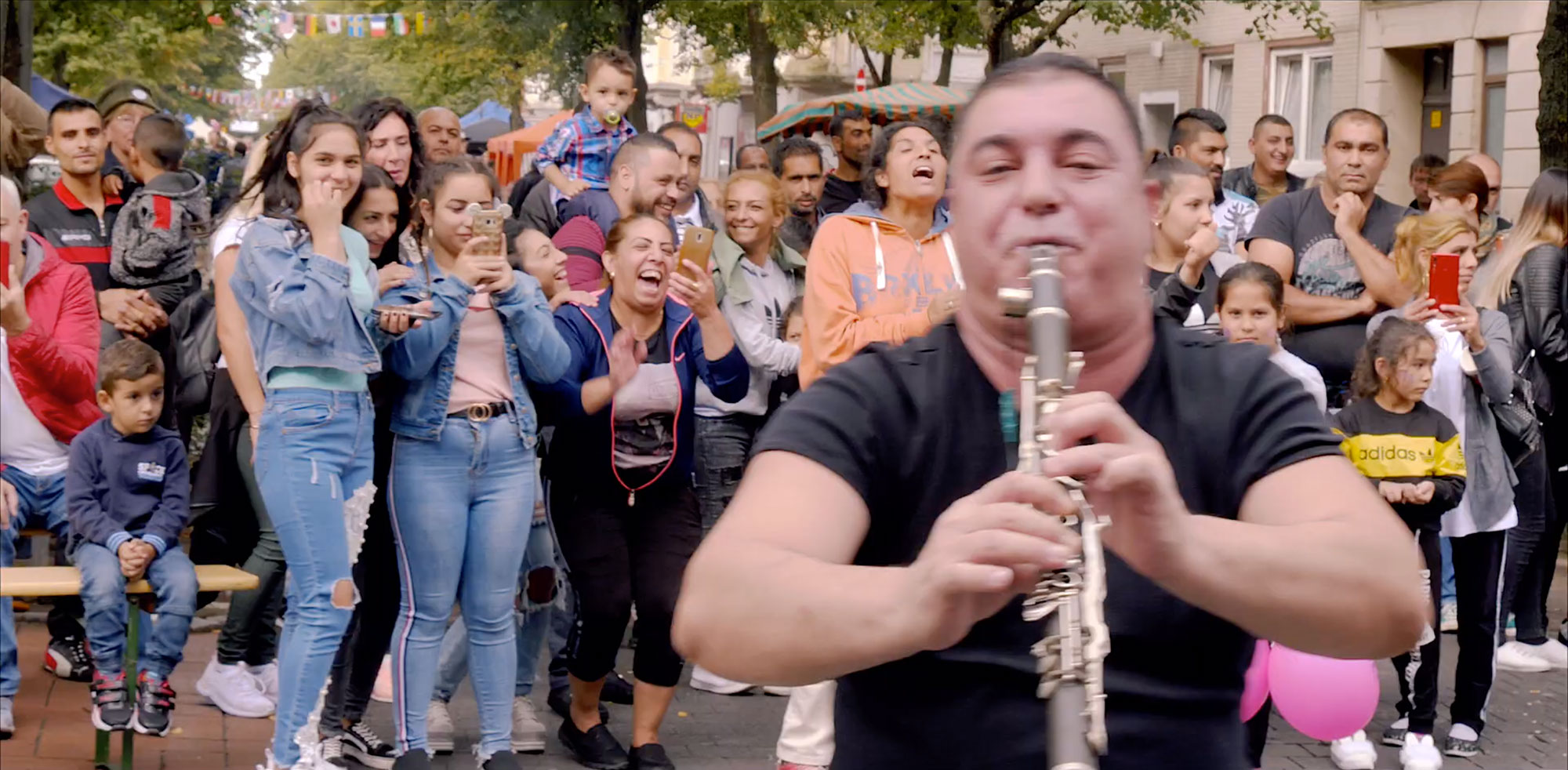 Menschenmenge steht hinter einem, im Vordergrund zu sehenden, Musiker. Der Musiker spielt eine Klarinette auf dem Goethestraßenfest. Die Straße ist mit Wimpelketten in den Bäumen geschmückt und im Hintergrund sind verschiedene Stände zu sehen.