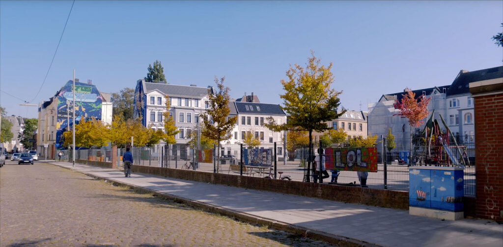 Blick auf den Leher Pausenhof. Hinter einem Zaun sind Spielgerüste, -geräte, Fahrräder und Kinder zu sehen.