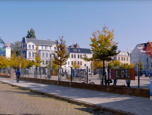 Blick auf den Leher Pausenhof. Hinter einem Zaun sind Spielgerüste, -geräte, Fahrräder und Kinder zu sehen.