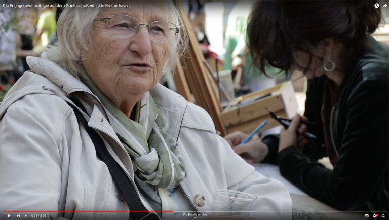 Videovorschaubild für das Goethestraßenfest. Ein ältere Frau mit einer Brille spricht. Im Hintergrund sieht man eine dunkelhaarige Frau mit Ohrringen, die etwas zeichnet.