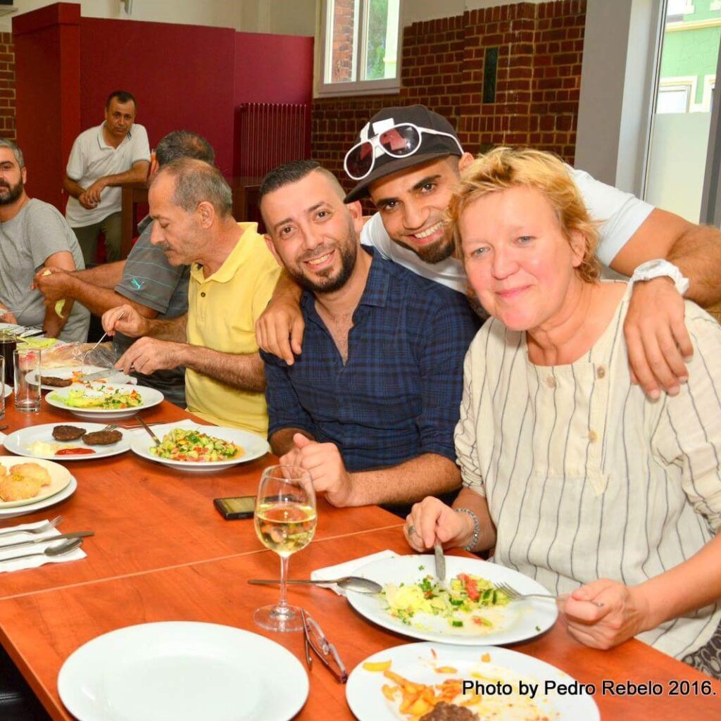 Bei dem Projekt „Esskulturen – Toleranz über den Tellerrand hinaus“ essen Menschen zusammen an einem langen Tisch. Im Vordergrund umarmt eine Person zwei weitere Personen von hinten. Alle drei lächeln in die Kamera.