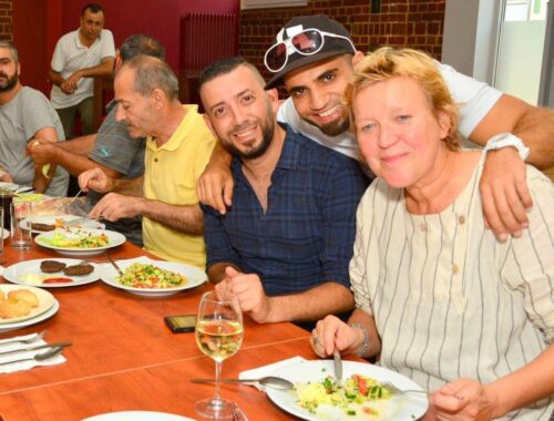 Bei dem Projekt „Esskulturen – Toleranz über den Tellerrand hinaus“ essen Menschen zusammen an einem langen Tisch. Im Vordergrund umarmt eine Person zwei weitere Personen von hinten. Alle drei lächeln in die Kamera.