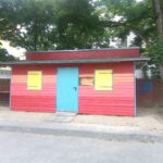 Die sogenannte „Bunte Bude“ vor einer Steinmauer des Leher Pausenhofs – eingerahmt von Bäumen. Die Bunte Bude verkauft Speisen und Getränke.