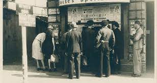 Schwarz/Weiß-Foto zeigt Menschen vor einem Kiosk stehend und interessiert die Zeitungsartikel lesend.