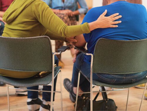 Menschen sitzen auf Stühlen im Kreis und eine Person legt trösten die Hand auf die Schulter der Person neben ihr