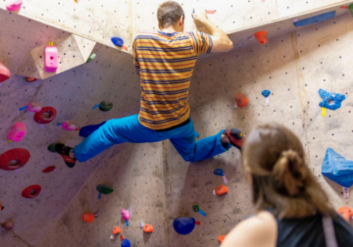 Männliche Person bouldert an einer Boulderwand. Im Vordergrund befindet sich eine weibliche Person, die ihm dabei zuschaut.