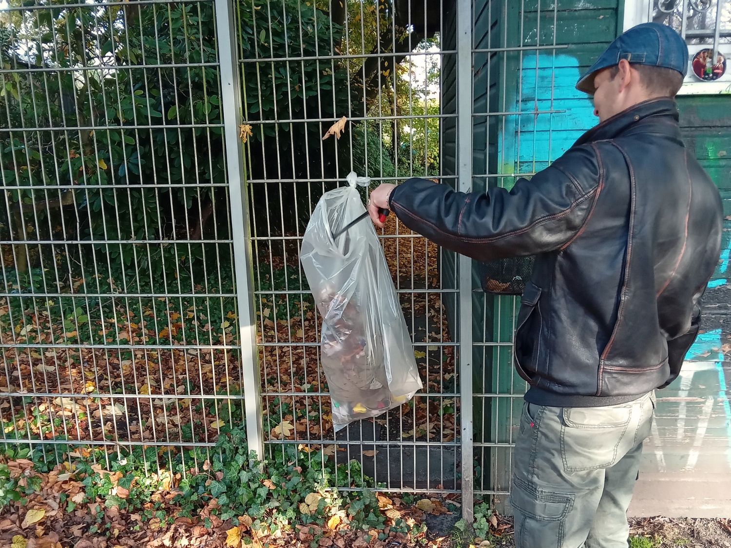 Mann sammelt mit einem Müllgreifer Müll auf und steckt ihn gerade in einen Müllsack, der an einem Metallzaun angebracht wurde.