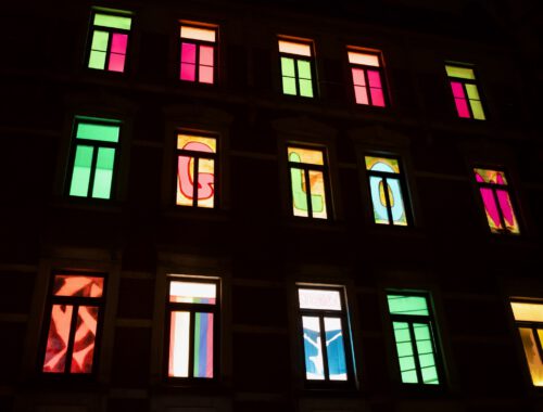 Fensterfront eines mehrstöckigen Altbauhauses in der Dunkelheit. Nur die beleuchteten Fenster sind zu erkennen. In jedem Fenster befindet sich eine andere bunte Gestaltung. In einer Reihe ergeben einzelne Buchstaben je Fenster, das Wort GLOW.