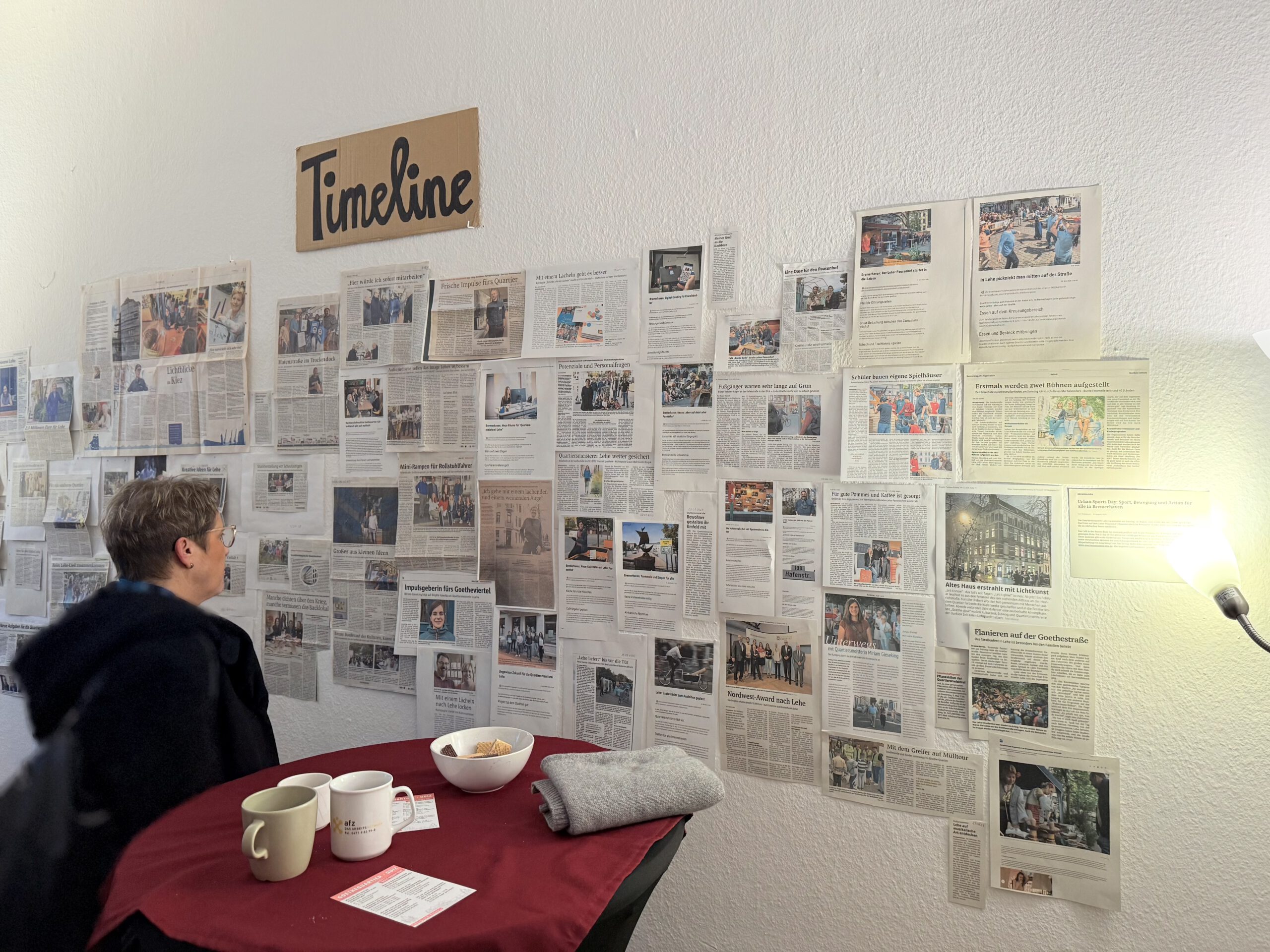 Frau mittleren Alters steht vor einer Wand, mit vielen Zeitungsartikeln und guckt sich diese an.