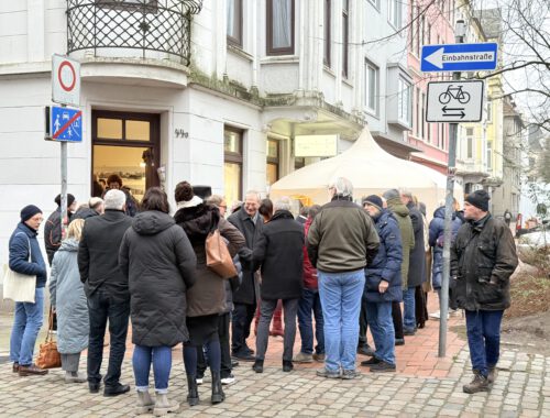 Viele Menschen stehen auf dem Bürgersteig vor einem Eckhauseingang, dem EIngang zur Quartiersmeisterei. IM Hintergrund ist ein weißer Pavillon zu sehen.