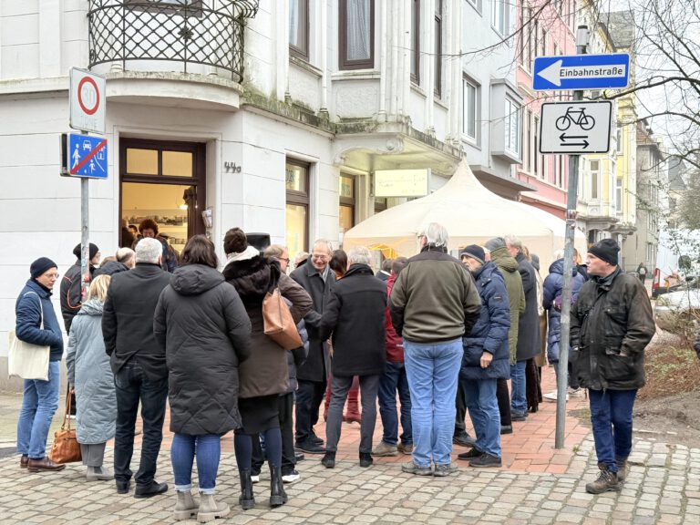 Viele Menschen stehen auf dem Bürgersteig vor einem Eckhauseingang, dem EIngang zur Quartiersmeisterei. IM Hintergrund ist ein weißer Pavillon zu sehen.