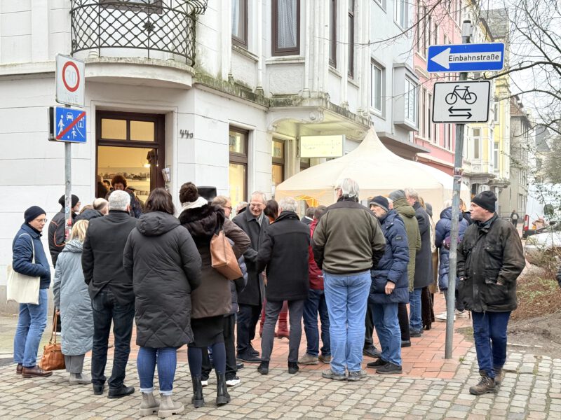 Viele Menschen stehen auf dem Bürgersteig vor einem Eckhauseingang, dem EIngang zur Quartiersmeisterei. IM Hintergrund ist ein weißer Pavillon zu sehen.