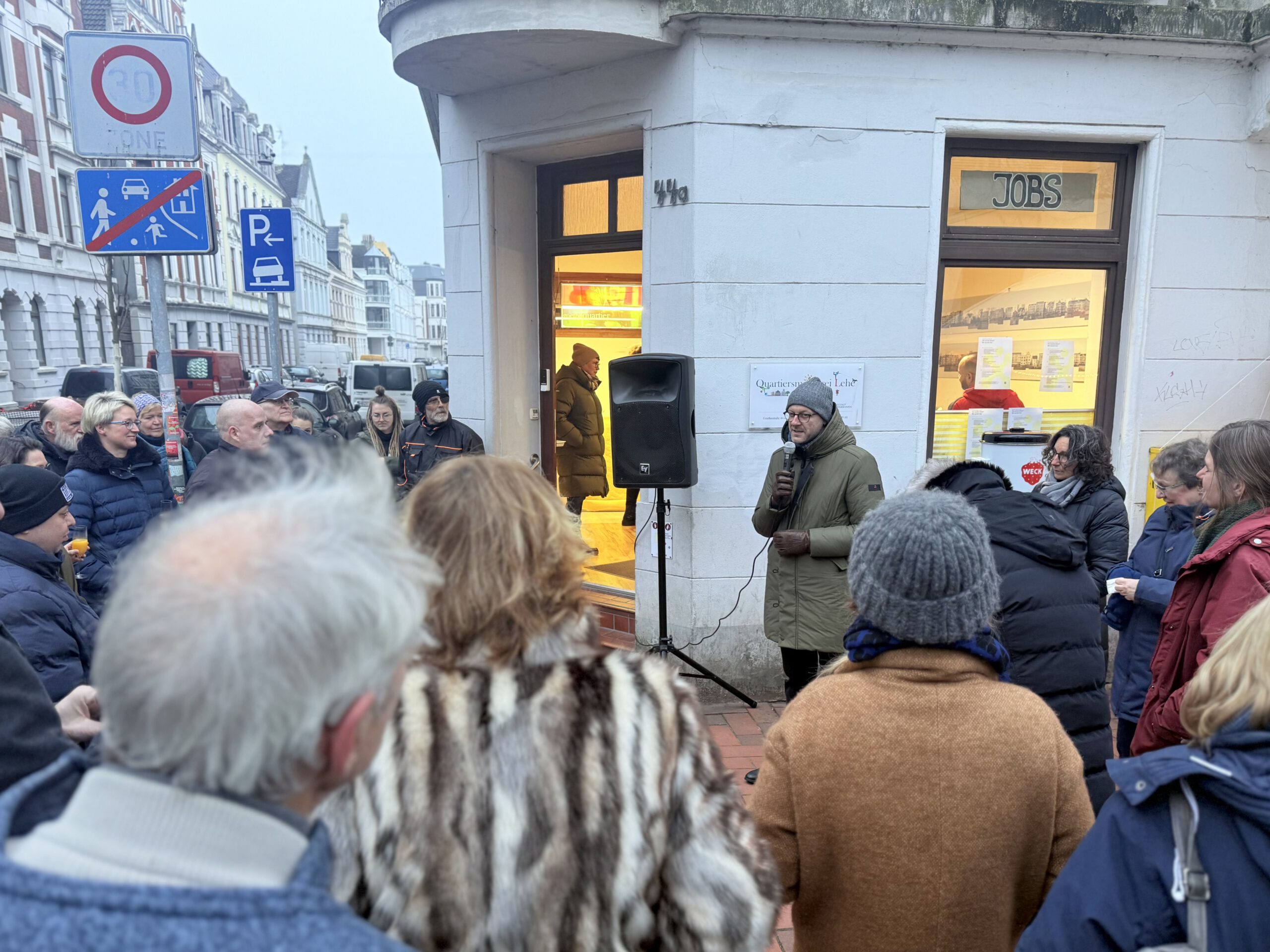 Menschenmenge vor einem weißen Eckhaus. Alle Blicke sind auf einen Mann, Stadtrat Martin Günthner gerichtet, welcher eine Rede hält.