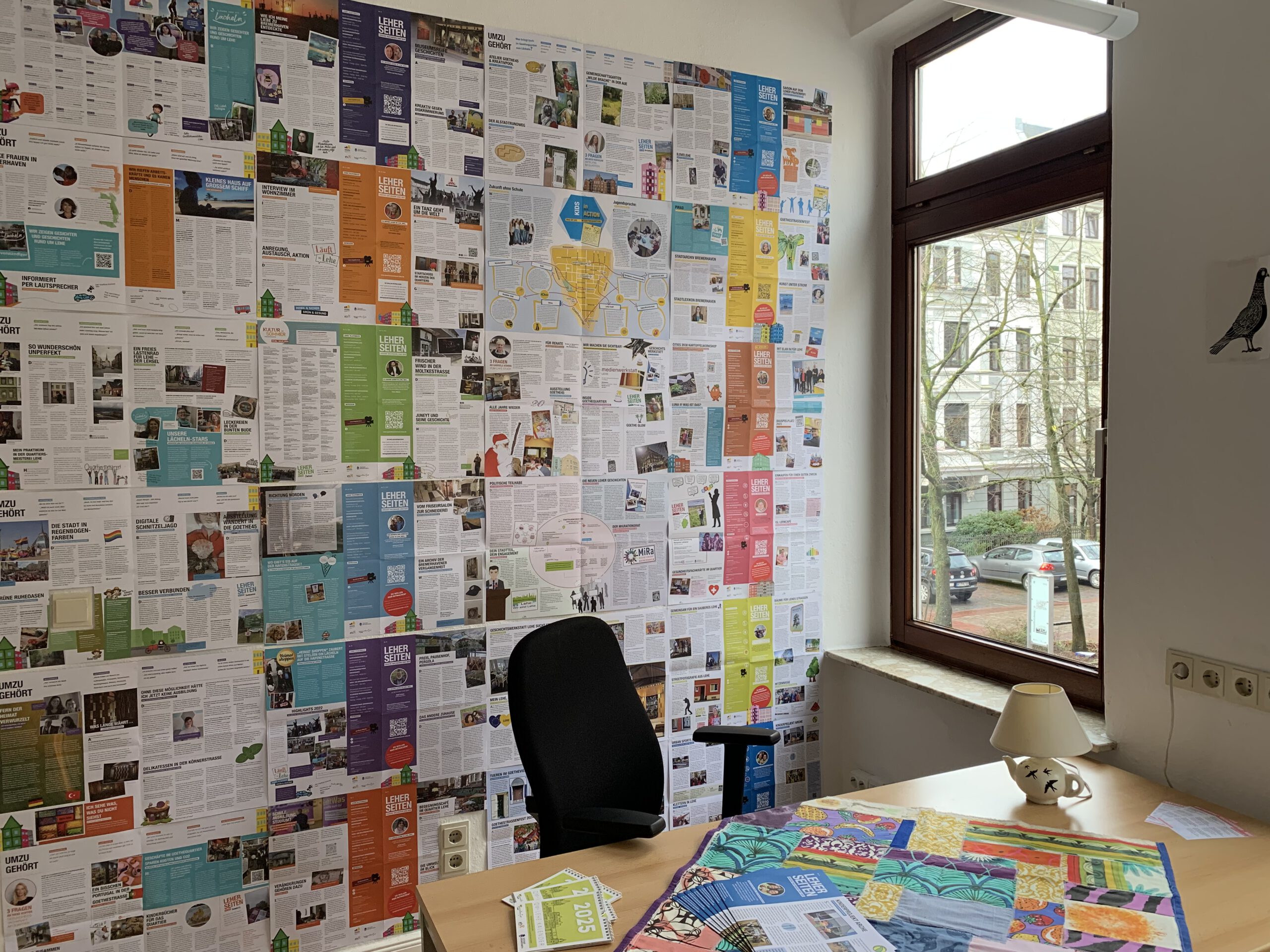 An der Wand eines Büros hängen ineinander übergehend bunte Ausgaben der Stadtteilzeitung 
