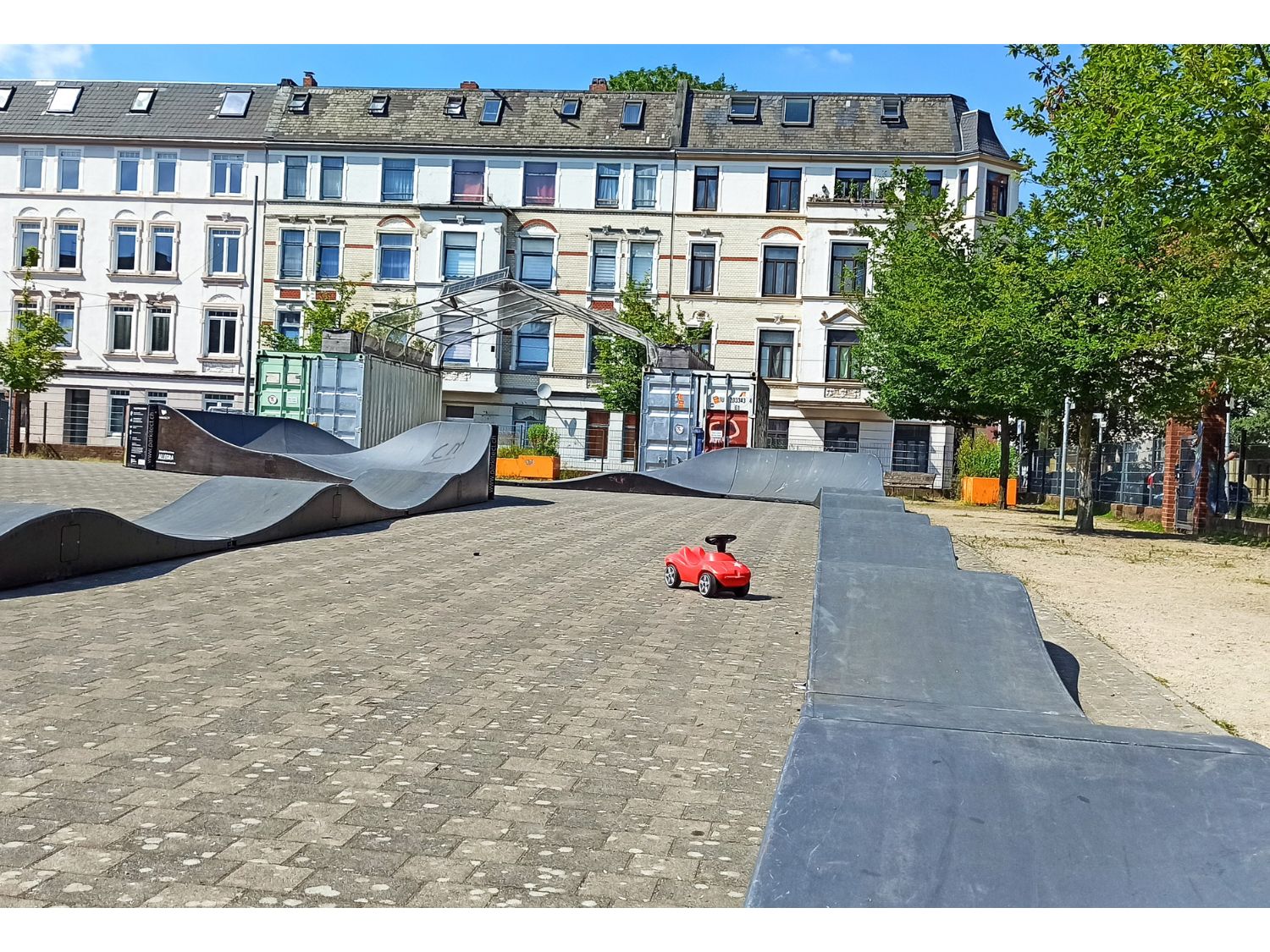 Gepflasterter Platz mit einem mobilen Pumptrack und einem roten Bobbycar. Im Hintergrund ist die Fassade eines Altbaus zu sehen.