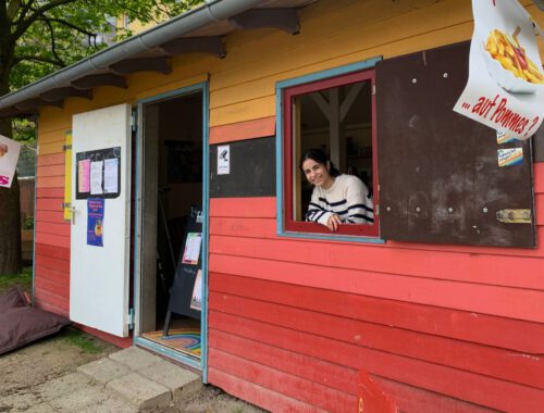 Eine bunte Holzbude mit einer jungen weiblich gelesenen Person die aus dem Fenster schaut