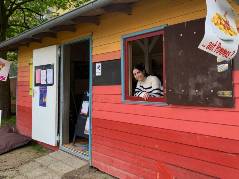 Eine bunte Holzbude mit einer jungen weiblich gelesenen Person die aus dem Fenster schaut