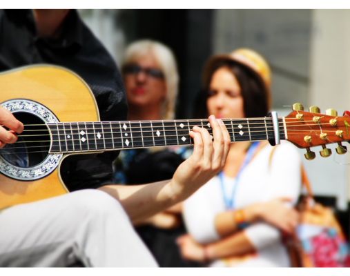 Nahaufnahme einer Gitarre, die gespielt wird. Im Hintergrund laufen verschwommen zwei Frauen vorbei.
