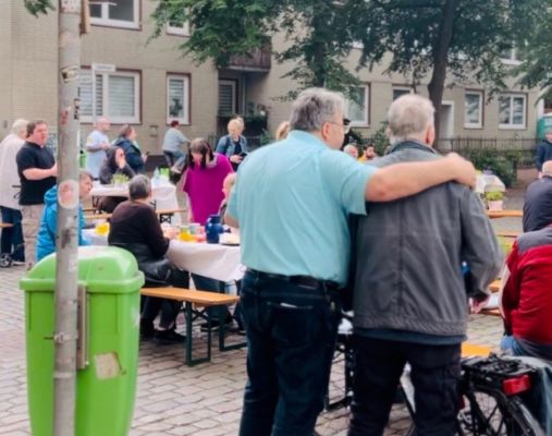 Bierzeltbänke und -tische sind auf einer Straßenkreuzung aufgestellt. Viele Menschen sitzen und stehen dort. Im Vordergrund legt ein Mann den Arm um einen anderen Mann