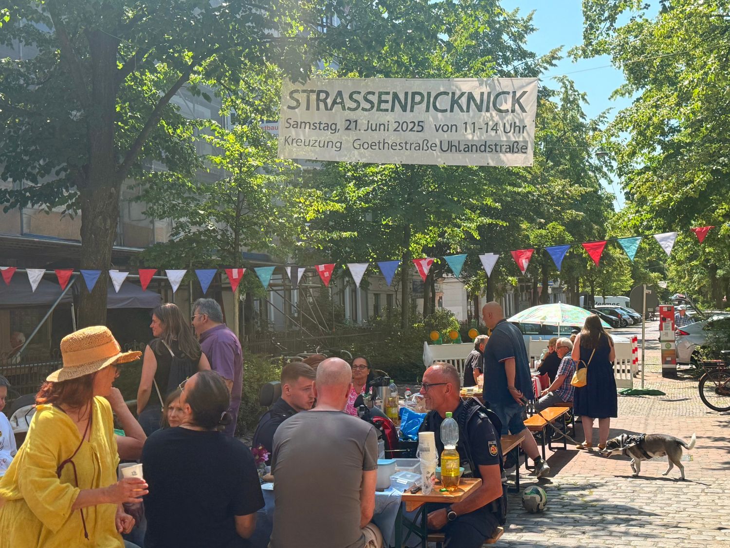 Viele Menschen sitzen an Tischen auf einer Straßenkreuzung unter dem Banner 