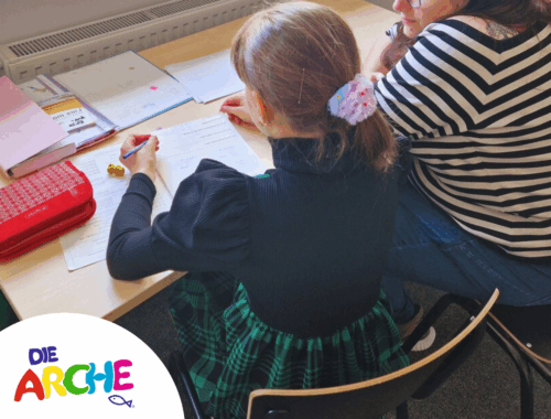 Ein Kind sitzt an einem Tisch und eine erwachsene Person daneben