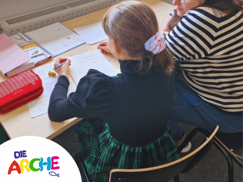 Ein Kind sitzt an einem Tisch und eine erwachsene Person daneben