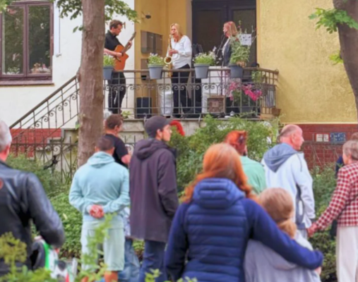 Mehrere Menschen stehen auf der Straße und schauen Musiker*innen in einer Eingangstür einer alten Villa zu