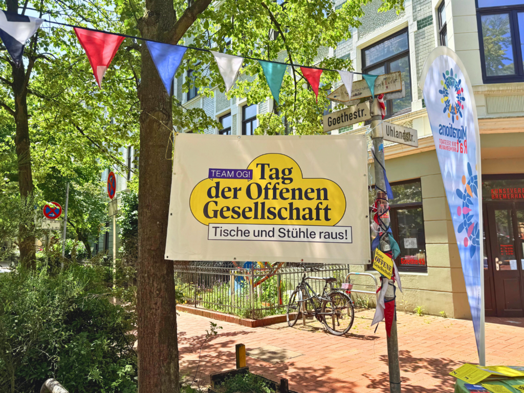 Ein Banner vom Tag der offenen Gesellschaft hängt zwischen einem Baum und einem Straßenschild. es ist Sommer auf der Goethestraße
