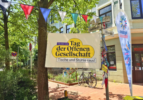 Ein Banner vom Tag der offenen Gesellschaft hängt zwischen einem Baum und einem Straßenschild. es ist Sommer auf der Goethestraße