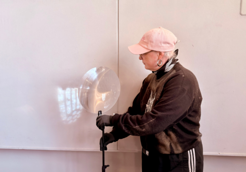 Eine Person hält ein Stativ mit einer Lichtreflektierenden Linse vor eine weißen Wand auf die ein umgedrehtes Kirchenfenster projiziert wird