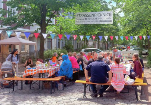 viele Menschen sitzen an Tischen auf einer Straßenkreuzung, über ihnen hängt eine bunte Wimpelkette und ein Banner auf dem 
