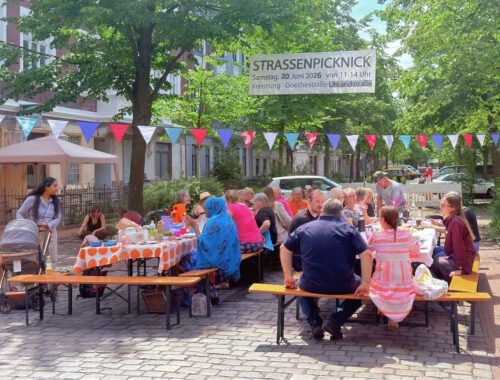 viele Menschen sitzen an Tischen auf einer Straßenkreuzung, über ihnen hängt eine bunte Wimpelkette und ein Banner auf dem 